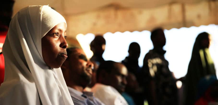 At night, a young African woman waring a white Khimar is standing in the left hand corner of the image and looks to something invisible towards the right hand corner of the image.